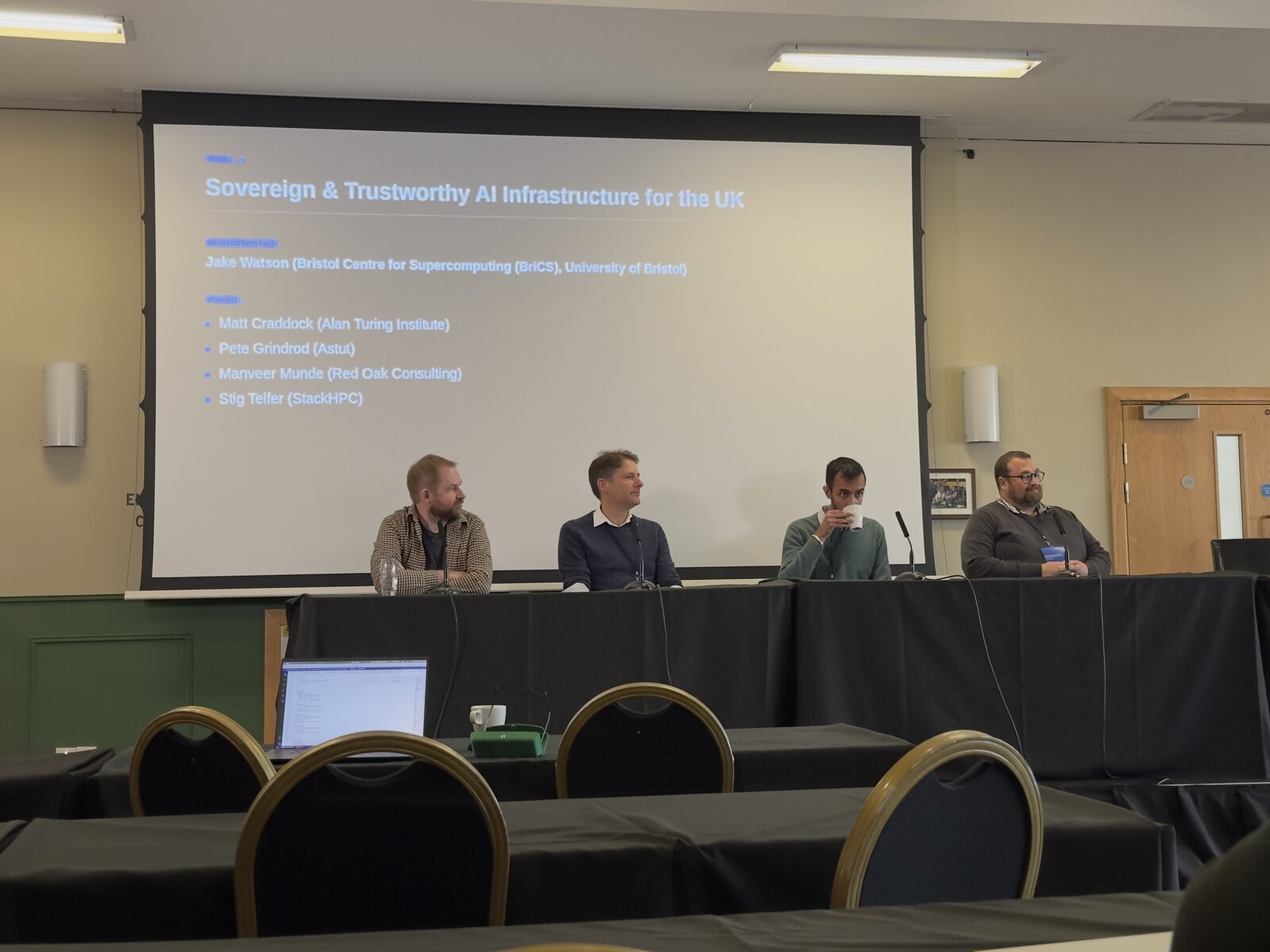 Photo of the panel: four panellists at a table under the slide listing the moderator, Jake Watson (Bristol Centre for Supercomputing (BriCS), University of Bristol), and the panel, Matt Craddock (Alan Turing Institute), Pete Grindrod (Astut), Manveer Munde (Red Oak Consulting) and Stig Telfer (StackHPC).