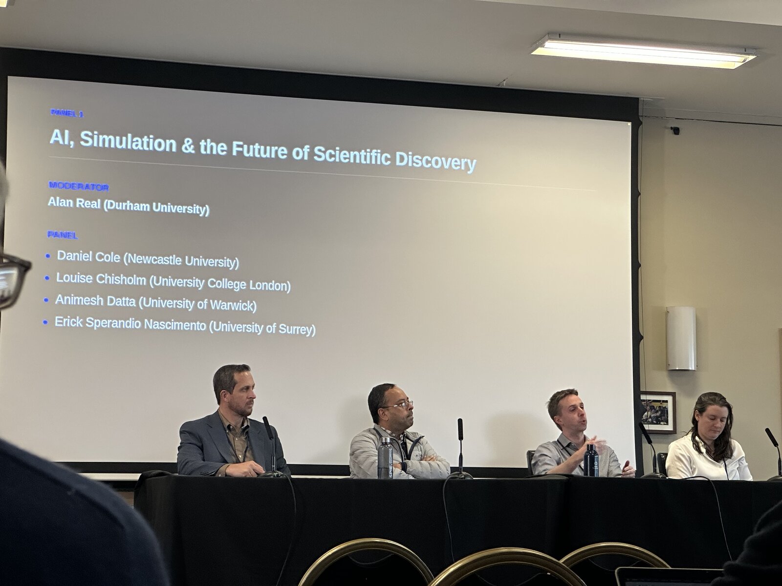 Photo of the panel: four panellists at a table under the slide listing the moderator, Alan Real (Durham University), and the panel, Daniel Cole (Newcastle University), Louise Chisholm (University College London), Animesh Datta (University of Warwick) and Erick Sperandio Nascimento (University of Surrey).